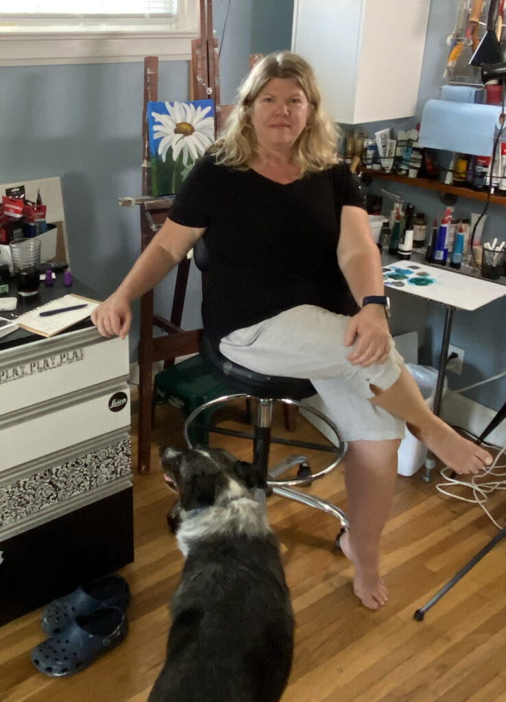 Artist Robin Nisbet sitting with her back to her easel. There is a medium sized dog in the foreground.
